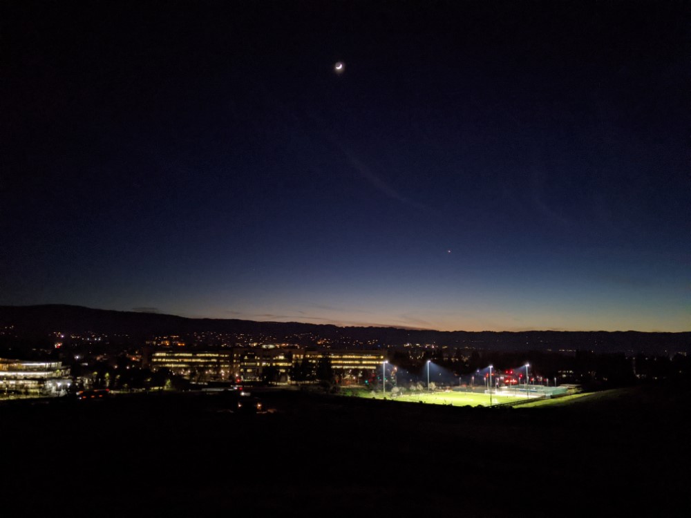 Picture overlooking Google's campus at night. An athletic field is in the foreground, and some short offices are behind it. In the distance, the sun has set behind some low mountains.